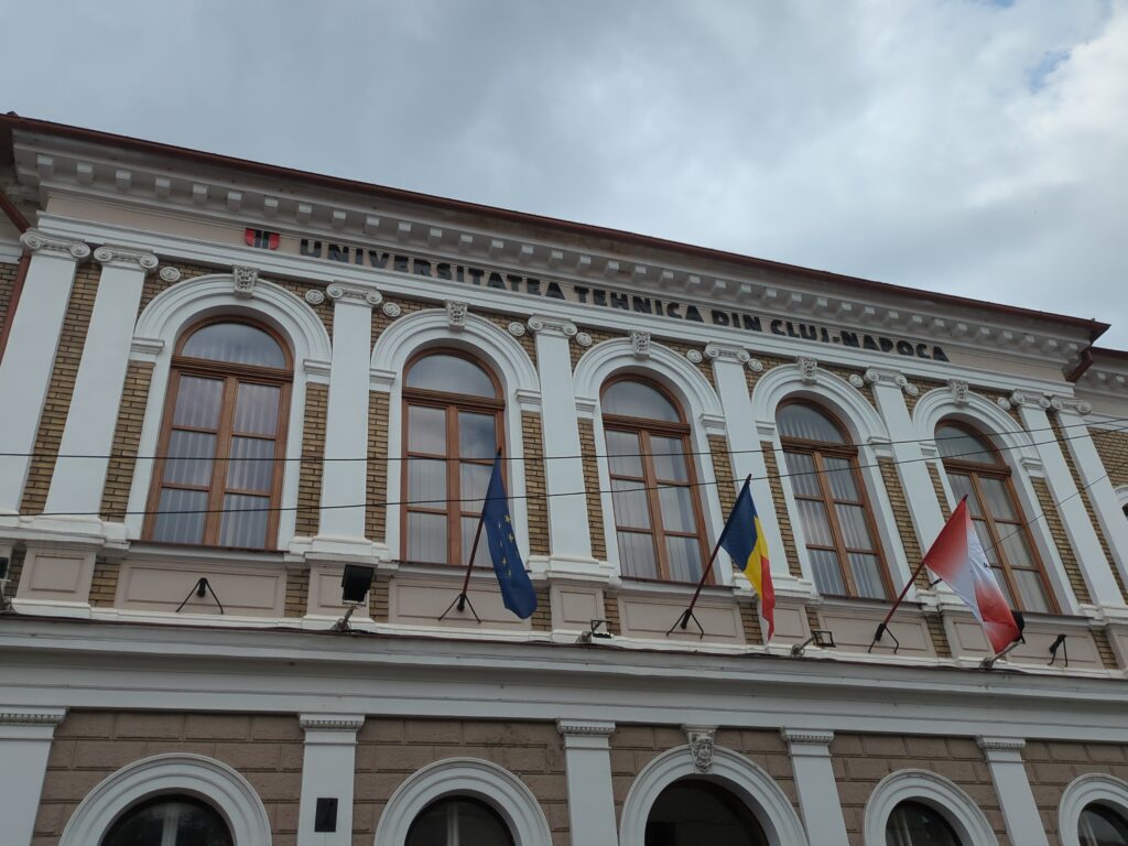 A historic building with arched windows and classical architectural details, displaying the name "Universitatea Tehnică din Cluj-Napoca" (Technical University of Cluj-Napoca) on the facade. Three flags are visible: the European Union flag, the Romanian national flag, and another flag, possibly representing a partner institution or regional symbol. The sky is overcast.
