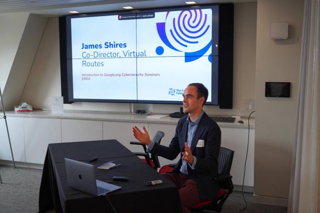 A man, identified on the screen behind him as James Shires, Co-Director of Virtual Routes, is speaking during a seminar. He is seated at a table with a black tablecloth, a laptop, and some electronic devices in front of him. Behind him, a large screen displays a slide titled “Introduction to Google.org Cybersecurity Seminars EMEA.” He gestures with his hands as he speaks, appearing engaged and expressive. The setting is a modern, well-lit conference room.